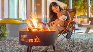 En person sitter vid en eldstad tillsammans med en hund, utomhus med en byggnad i bakgrunden.
