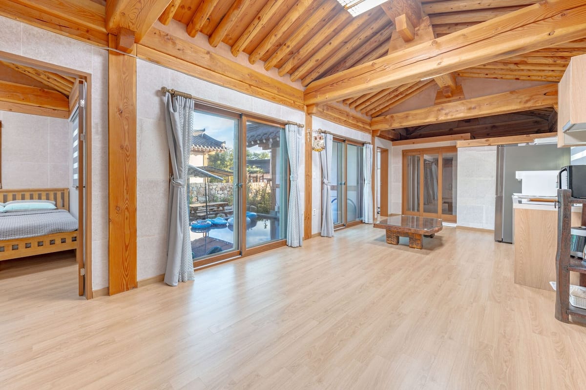 A spacious room with a wooden ceiling, a bed, a table, and a view of a pool and mountains through large sliding doors.