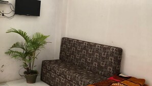 A small, simple room with a patterned sofa, a potted plant, and folded blankets on the floor. - Hotel Siddhartha (Ujjain)