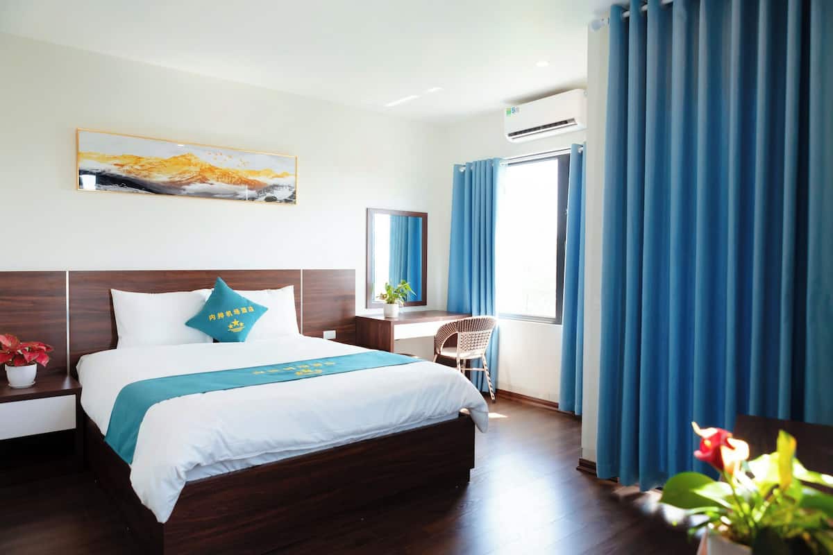 Ein Hotelzimmer mit einem großen Bett, einem Schreibtisch, einem Sessel, einer Topfpflanze und einem Vorhangfenster.