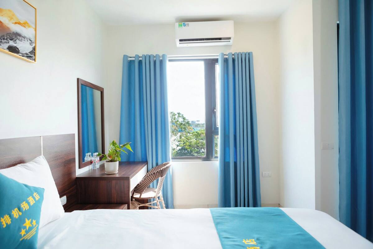 Ein Hotelzimmer mit Bett, Schreibtisch mit Stuhl, Fenster mit Vorhängen und einer Wand-Klimaanlage.