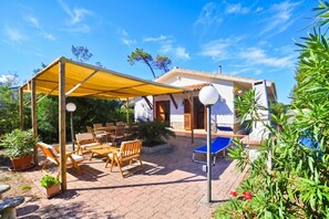 A covered outdoor seating area with wooden furniture, a blue lounge chair, and a white building with a tiled roof. - Villino Saline (Orbetello)