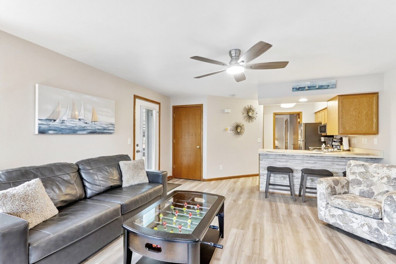 A living room with a sofa, coffee table, and a foosball table. There is a kitchen area with a bar and stools.