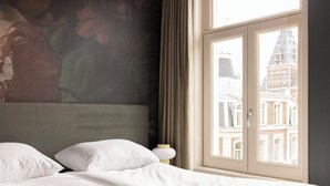A hotel room with a bed, a nightstand, and a view of a building through the window. - THE FRITZ HOTEL Amsterdam (Amsterdam)
