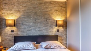 Une chambre à coucher moderne avec un mur à l’aspect pierre, deux lampes de chevet et un lit recouvert de linge de lit blanc.