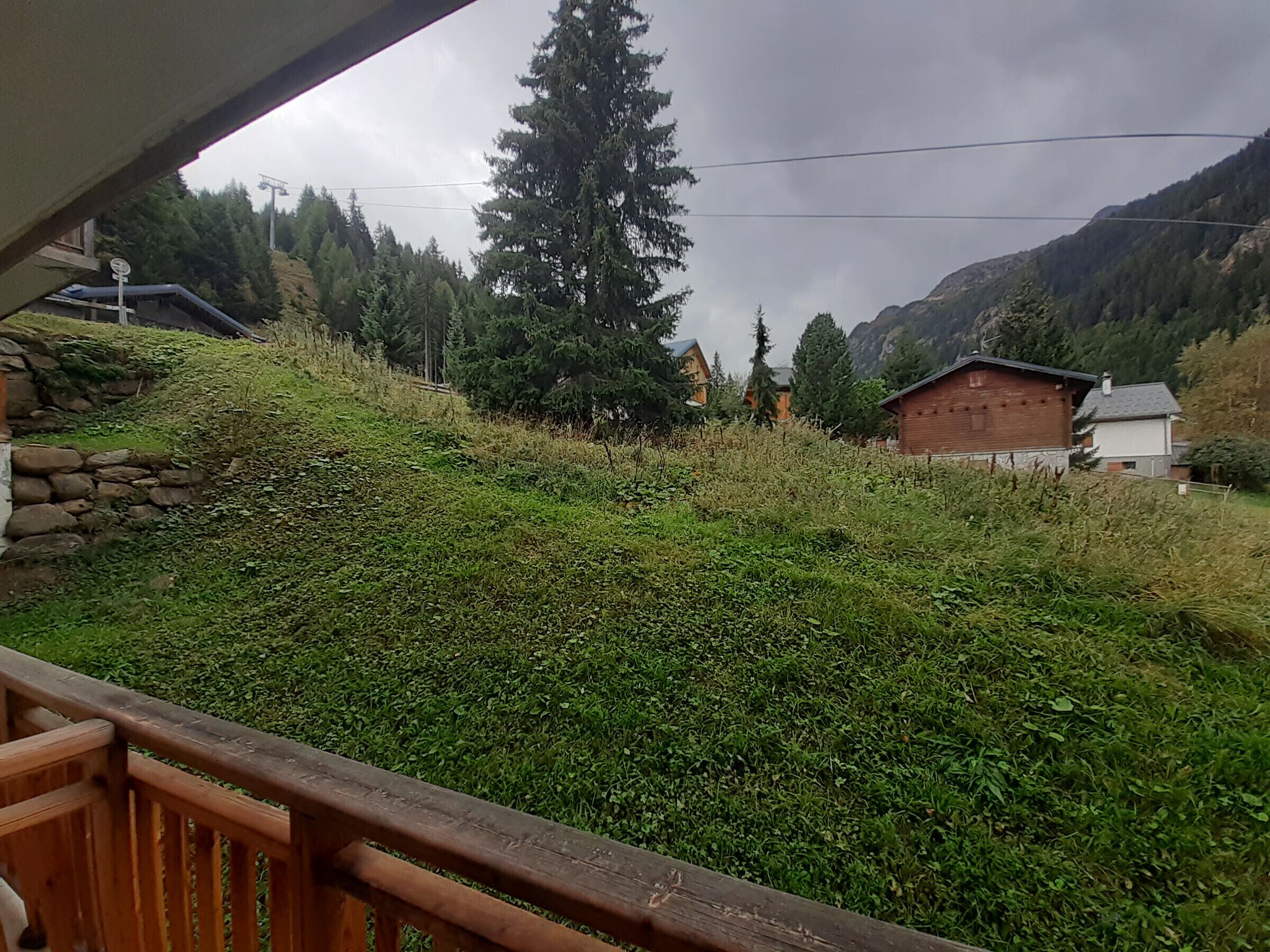 A mountainous landscape with a wooden deck overlooking a grassy area with a few buildings in the background.