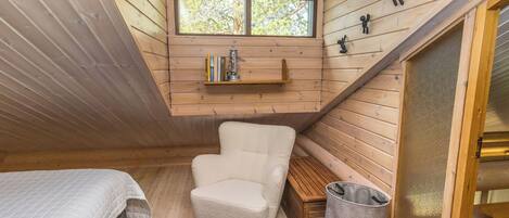 Une chambre mansardée chaleureuse avec un plafond en pente, une fenêtre, une bibliothèque, une chaise, un lit et une poubelle.