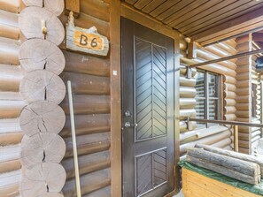 En hytte med træfacade og en trædør med skiltet 'B6', et vindue med et chevronmønster og en stabel brænde.