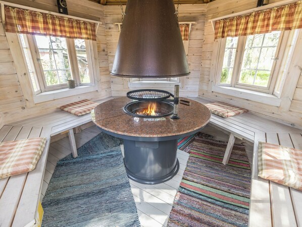 A circular stone fireplace with a metal hood, surrounded by wooden benches and a colorful woven rug.