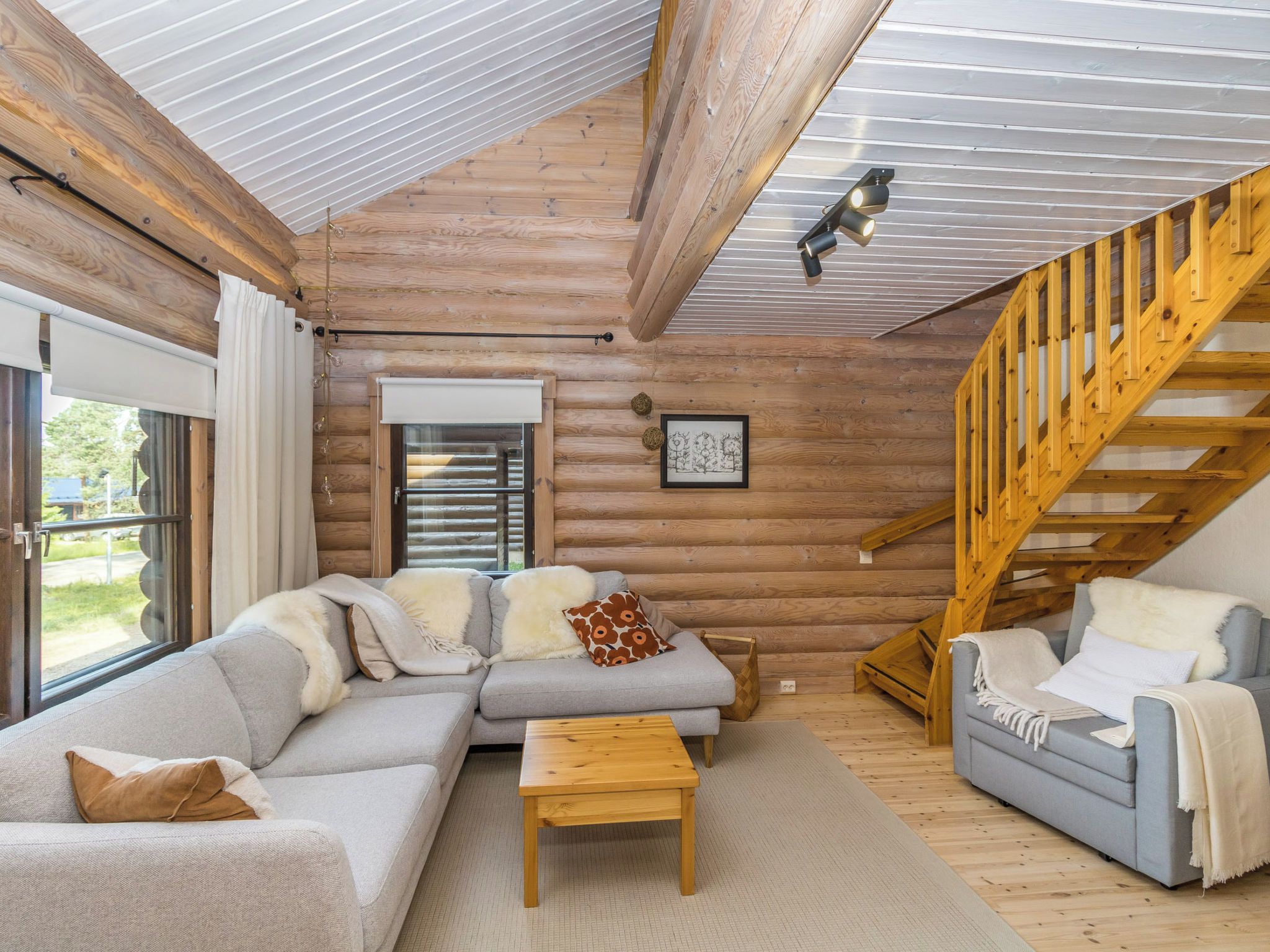 Interior de una cabaña de troncos con una escalera de madera, un conjunto de sofás y una mesa de comedor.