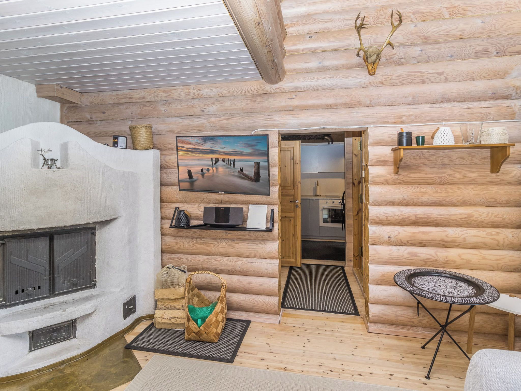 Interior de cabana de tronco com lareira, televisão na parede, espaço de cozinha e uma pequena mesa redonda.