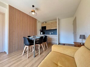 A modern living room with a yellow sofa, a dining area with a wooden wall, and a kitchen with wooden cabinets. - Cozy apartment at the foot of the slopes with pool (Flumet)