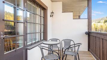 Um deck de madeira com mesa e cadeiras, uma porta com janelas amplas e vista para uma área arborizada.