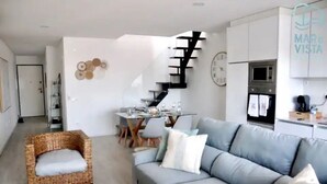 A modern living room with a grey sofa, a wicker chair, and a dining area with a table and chairs.