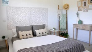 A bedroom with a large bed, a decorative headboard, a small round table with a plant, and a vanity with a mirror and a hat on the wall.