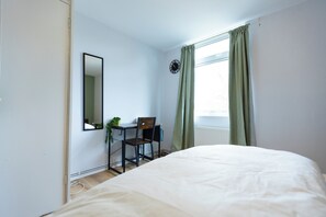 Une chambre à coucher avec un lit, un bureau, une chaise, une plante en pot et une fenêtre avec des rideaux.