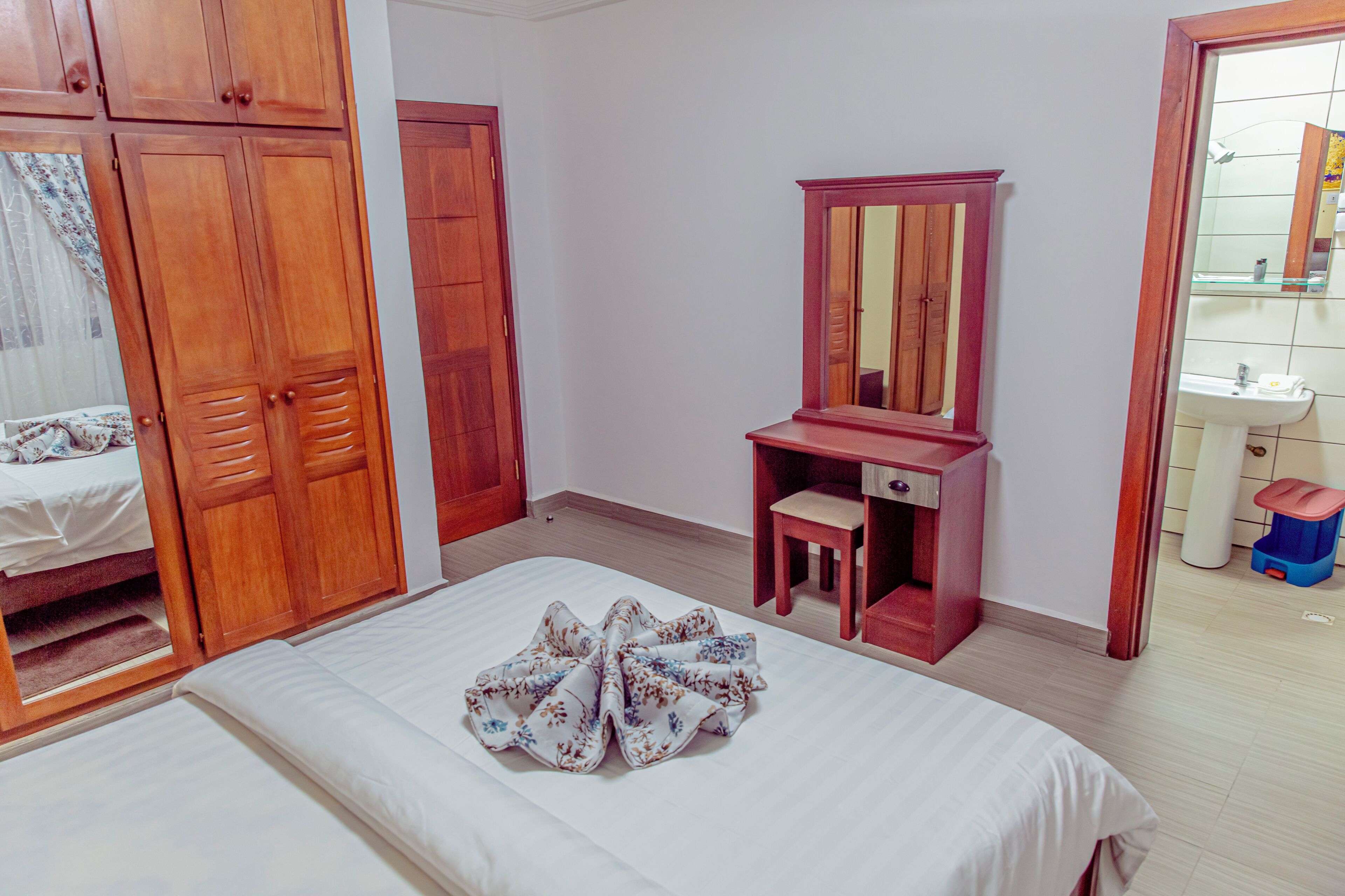 A bedroom with a bed, a wooden wardrobe, a desk with a mirror, and a bathroom visible through an open door.