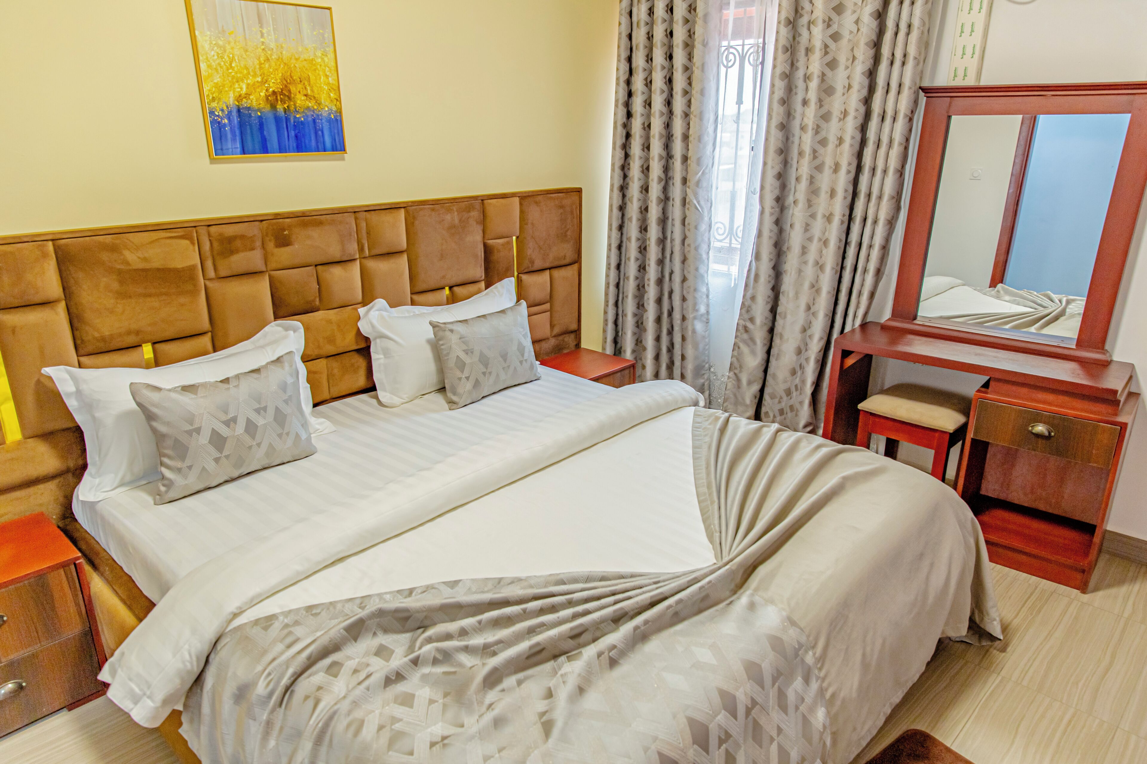 A neatly made bed with a headboard, a bedside table with a lamp, and a mirror on a wooden stand.
