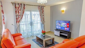 A living room with orange sofas, a coffee table with a vase, a TV on a stand, and a window with curtains. - G&G Residency (Douala)