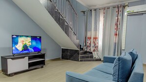 A living room with a TV, a staircase, a blue sofa, and a patterned curtain. - G&G Residency (Douala)