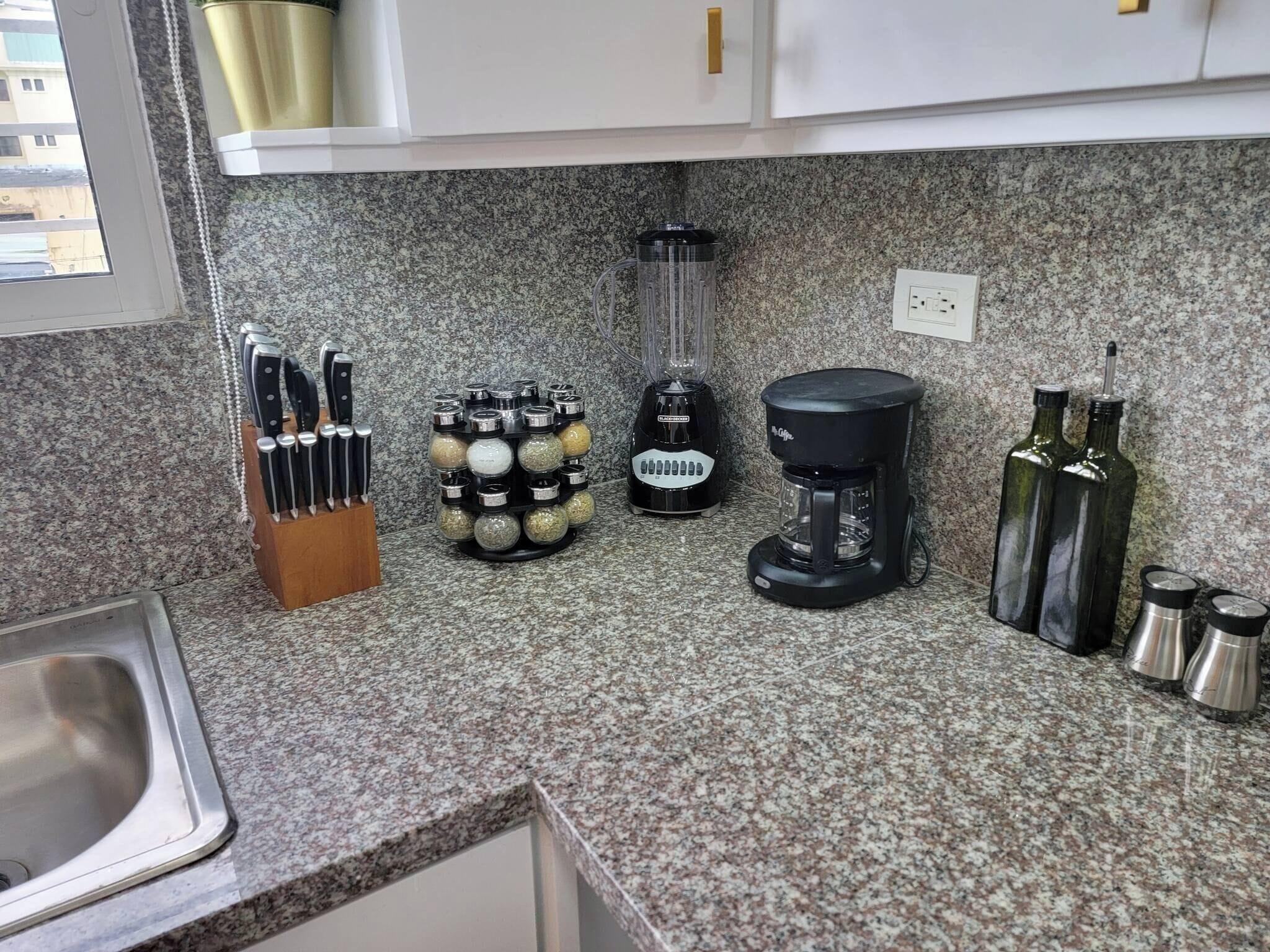 A kitchen with granite countertops, a knife block, spice jars, a blender, a coffee maker, and olive oil bottles.