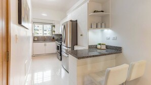 A modern kitchen with white cabinetry, a granite countertop, and stainless steel appliances. There is a small island with seating and a view of the outside. - Upscale 3 Br Apartment For Family Or Friends Stay (Santo Domingo)