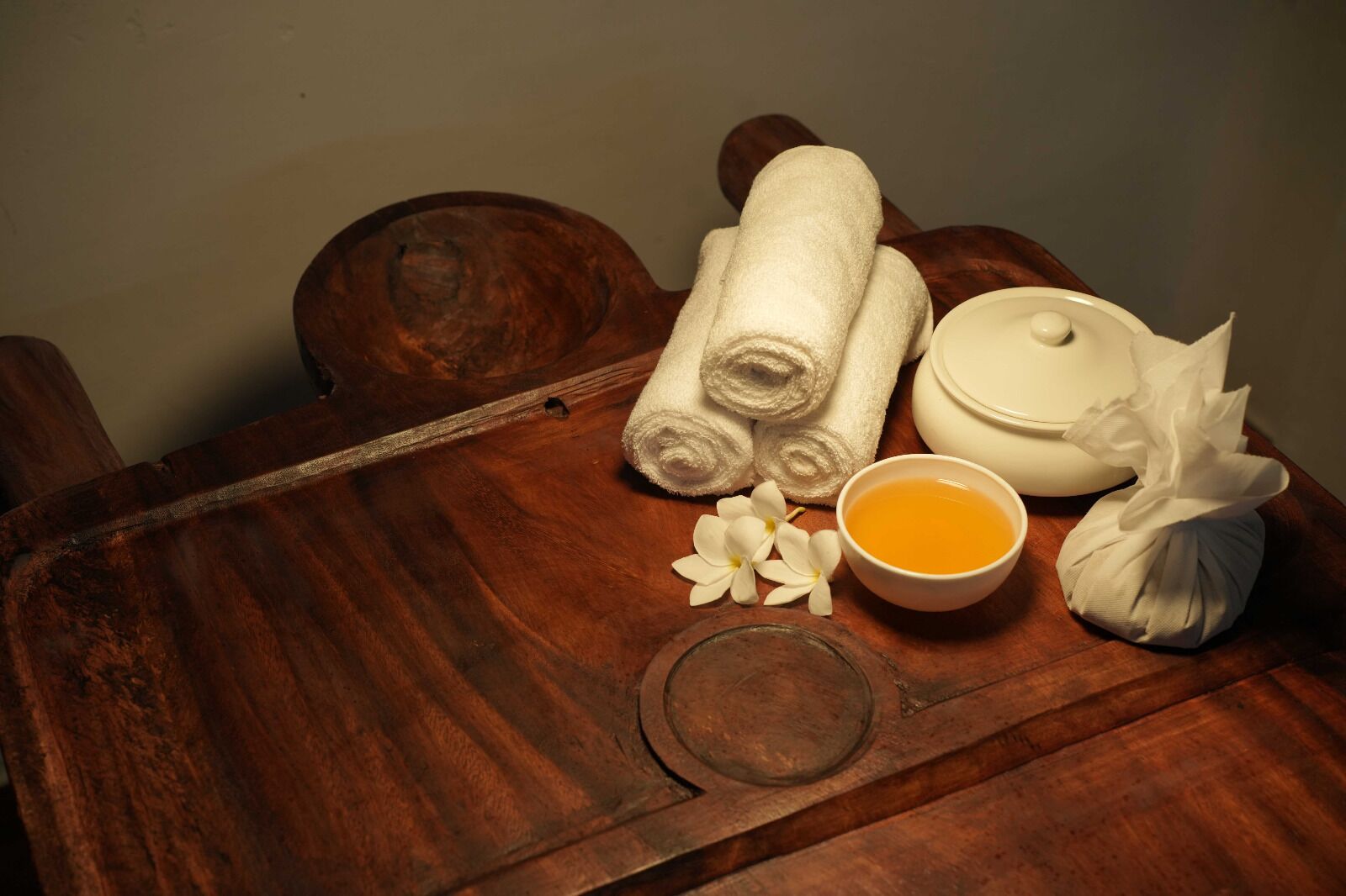 A wooden tray with rolled white towels, a white container, a bowl of yellow liquid, and a white cloth.