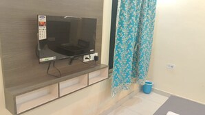 A flat-screen TV mounted on a wall with a control panel, a blue bucket, and a patterned curtain. - L S Grand By GRB (Varanasi)
