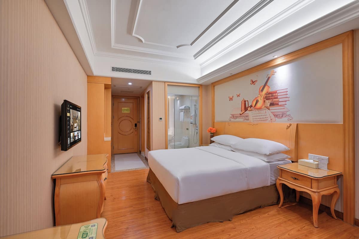 A hotel room with a large bed, a bedside table, a nightstand, a wall-mounted TV, and a door leading to a bathroom.