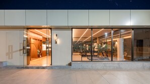 Ein modernes Gebäude mit großen Glasfenstern, einer Rezeption mit Holzvertäfelung und einem sichtbaren Fitnessraum mit Trainingsgeräten.