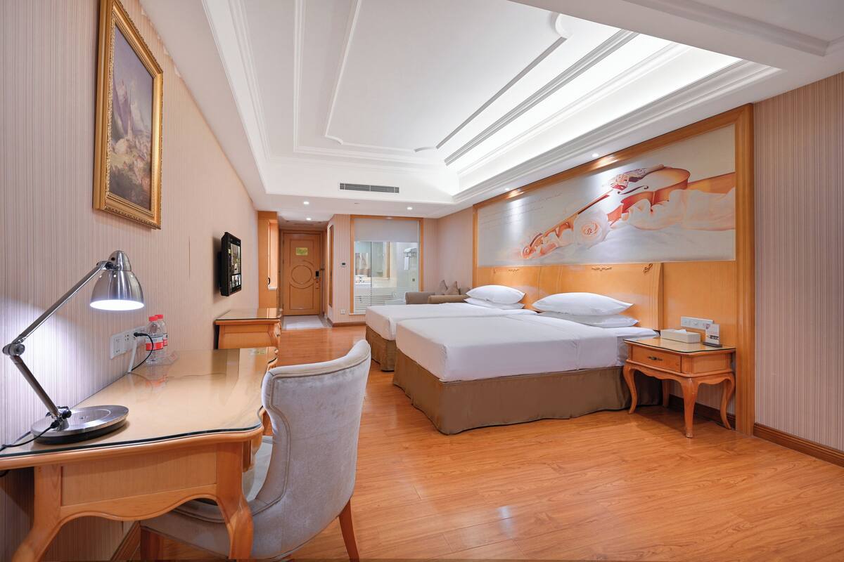 A hotel room with two beds, a desk with a lamp, a chair, a wooden nightstand, and a large painting on the wall.