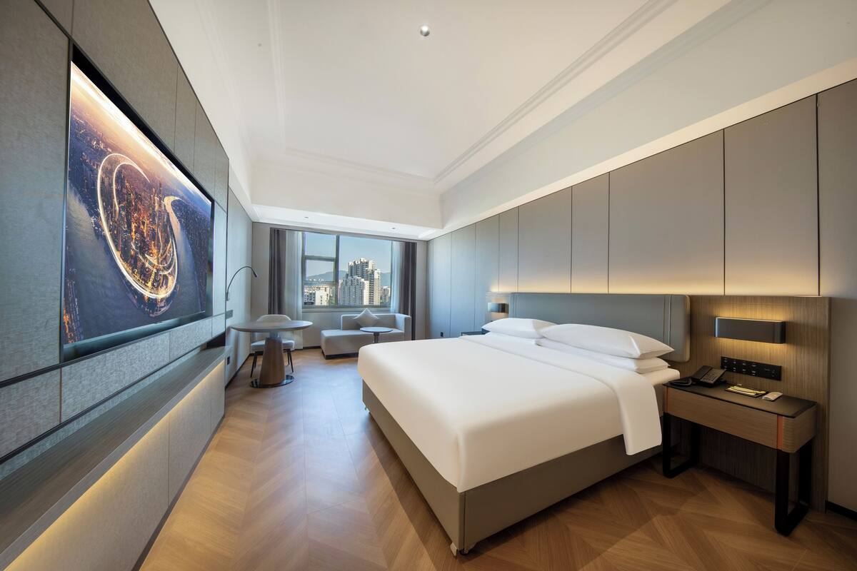 A modern hotel room with a large bed, a desk with a lamp, a small round table, and a large window offering a city view.