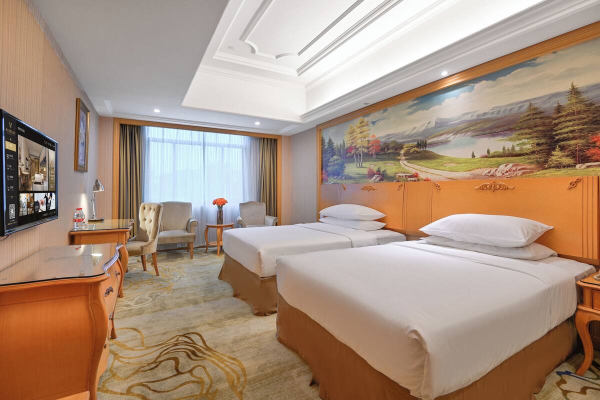 A hotel room with two beds, a desk, a chair, a TV, and a painting on the wall.