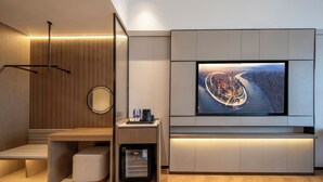 Ein modernes Hotelzimmer mit großem Flachbildfernseher an der Wand, einer Sitzecke mit einer Bank, einem kleinen Kühlschrank und einer Kaffeemaschine.
