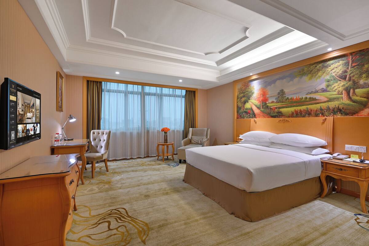 A hotel room with a large bed, a desk, a chair, a small table with a vase, and a painting on the wall.