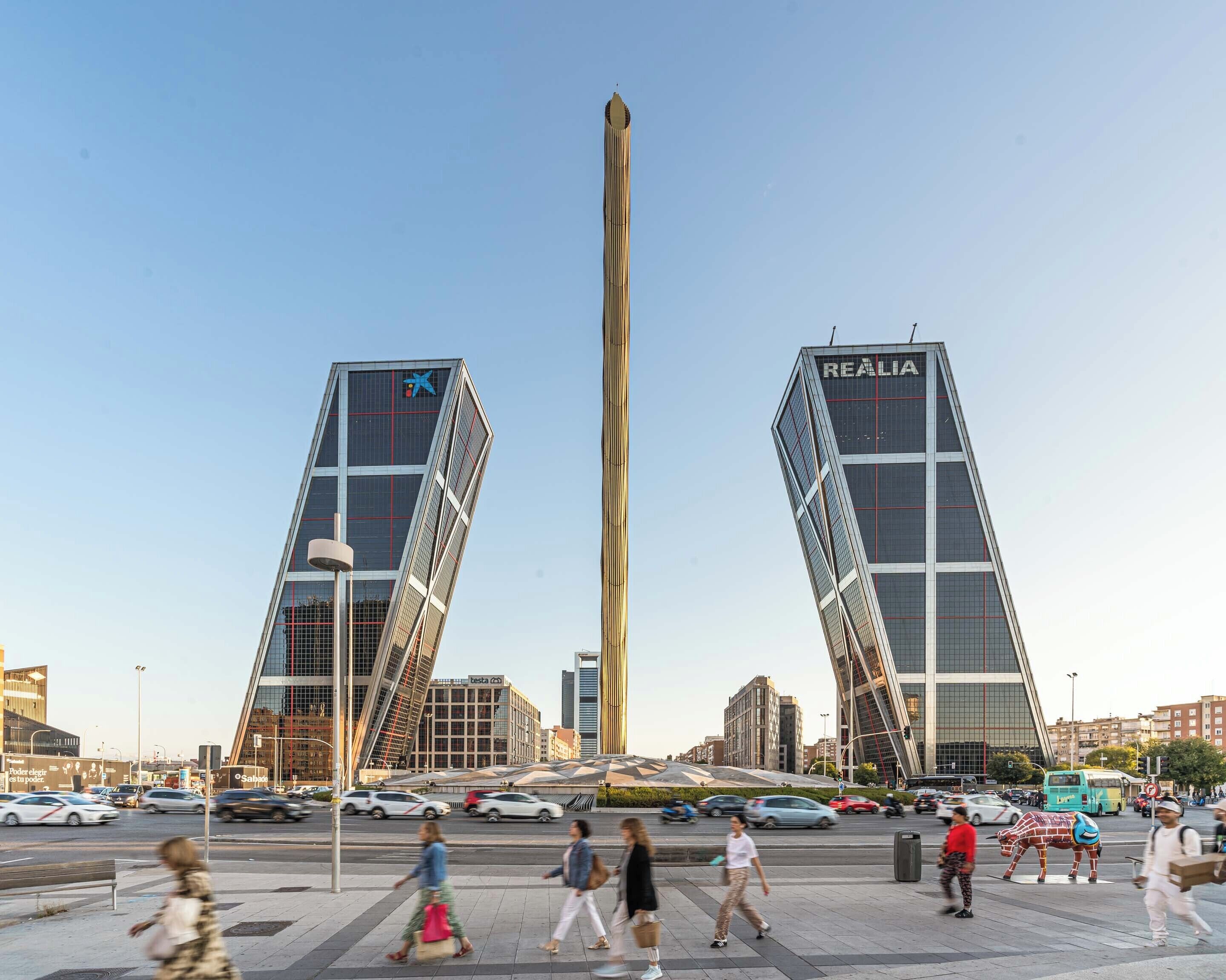 A modern urban square with two prominent buildings, a tall vertical structure, and a busy street with pedestrians and vehicles.
