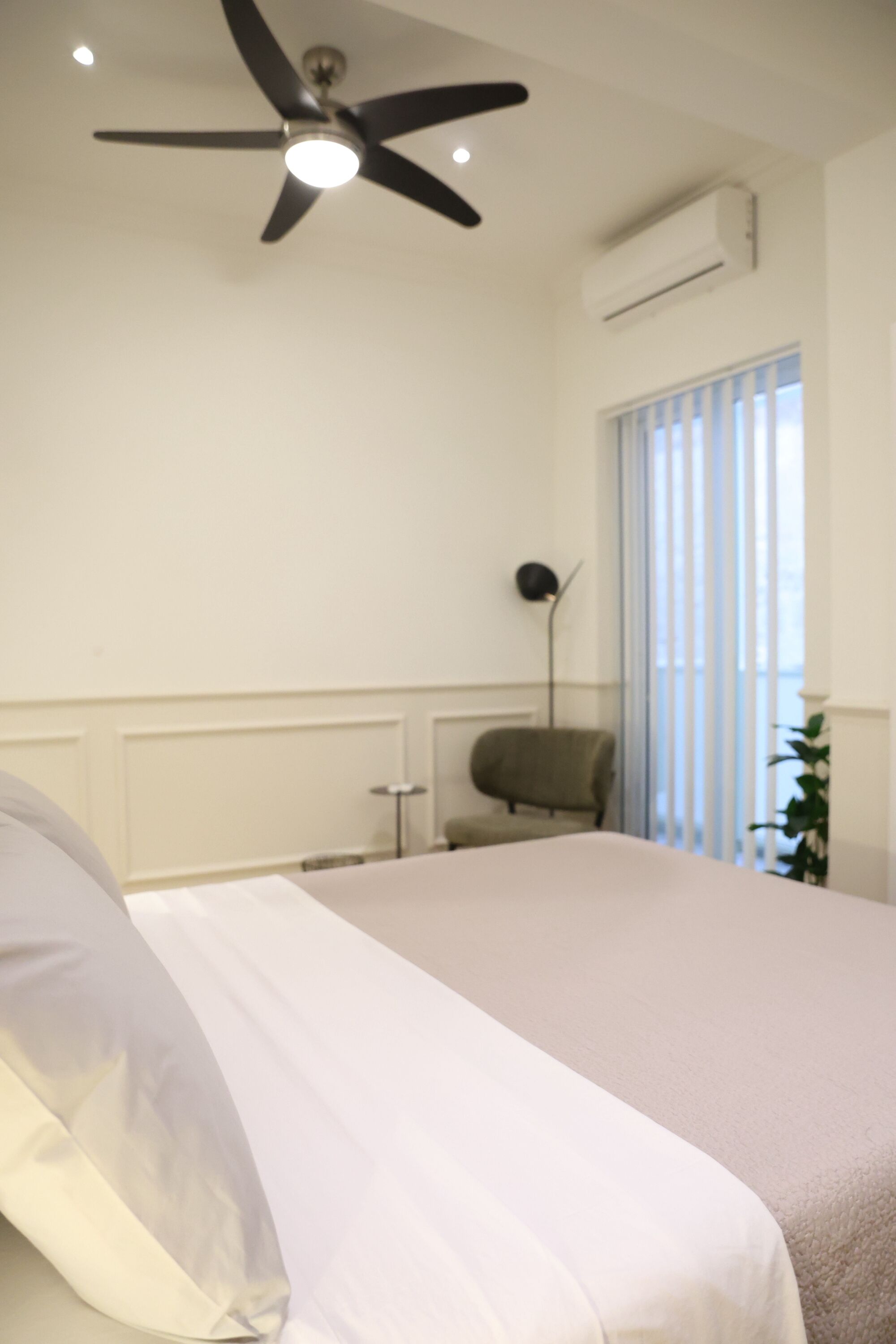 Une chambre à coucher comprenant un lit, un ventilateur de plafond, un appareil de climatisation, une chaise et une fenêtre avec des stores.
