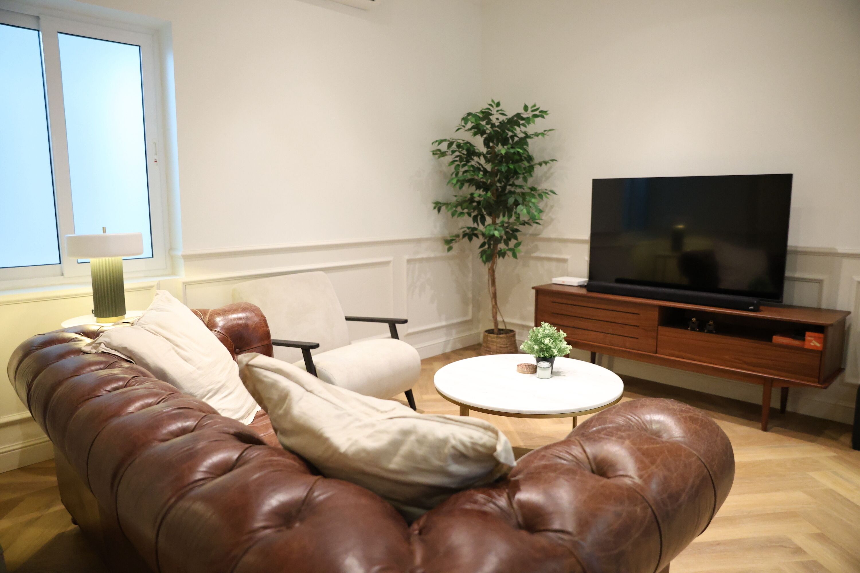 A living room with a brown leather sofa, a white armchair, a small round coffee table, a television on a wooden stand, and a potted plant.