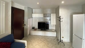 A compact living space with a kitchenette, a flat-screen TV, a coat rack, and a refrigerator.