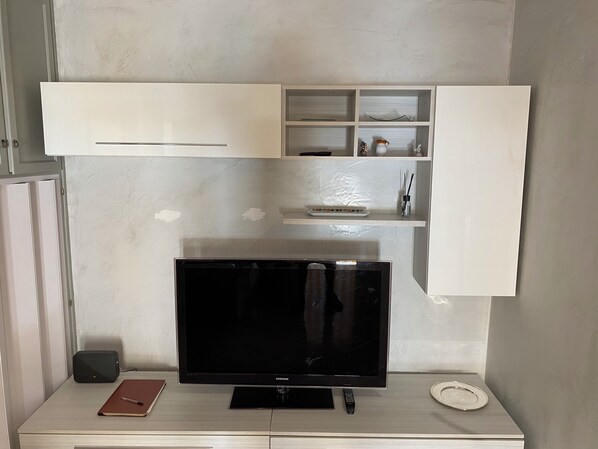 A modern living room with a flat-screen TV, a white cabinet with shelves, and a small table with a remote control.