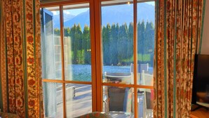 A room with a large window overlooking a lake and forest, with curtains and a television on the right. - Alpenhotel Rosenhof Ruhpolding (Ruhpolding)