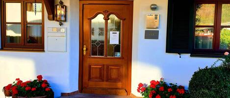 A wooden front door with a glass panel, a keypad lock, and a doorbell. There are flower pots with red flowers on either side of the door.