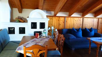 Una habitación acogedora con techo de madera, un sofá azul con cojines, una mesa de comedor de madera con sillas y una chimenea blanca con un arco decorativo.