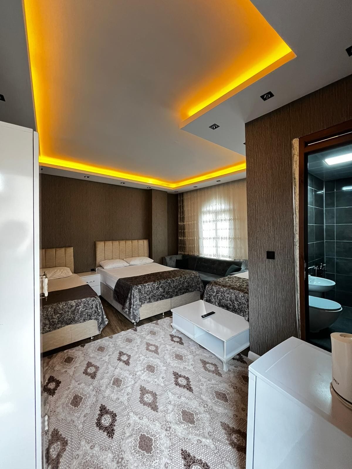 A modern hotel room with two beds, a sofa, a coffee table, and a bathroom with a sink and mirror.