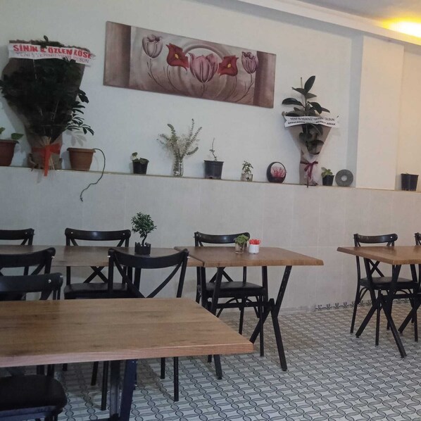 L’intérieur d’un restaurant est agrémenté de tables et de chaises en bois, d’un tableau représentant des fleurs accroché au mur et de diverses plantes en pot.