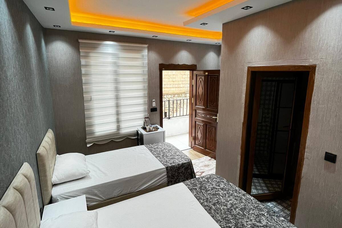 A hotel room with two beds, a small table, a window with blinds, a door, and a balcony visible through the window.
