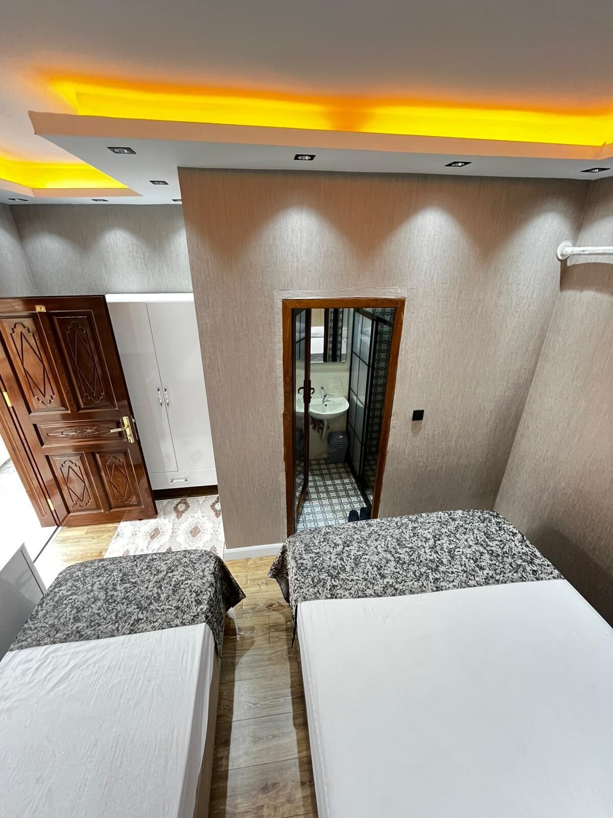 A hotel room with two beds, a wooden wardrobe, a bathroom visible through a doorway, and a ceiling with recessed lighting.