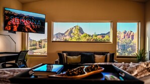 Habitación de hotel con una televisión grande mostrando 'SEDONA ARIZONA'S RED ROCK COUNTRY', una cama con una bandeja de aperitivos, un escritorio con una computadora portátil y vistas a formaciones rocosas rojas a través de dos ventanas.