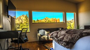 Un dormitorio con cama, escritorio y silla. Dos ventanas ofrecen vista a la montaña.