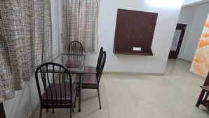 A dining area with a glass table and four chairs, a wooden cabinet, and a wall-mounted shelf. - PPH Living Arvind Inn (Coimbatore)